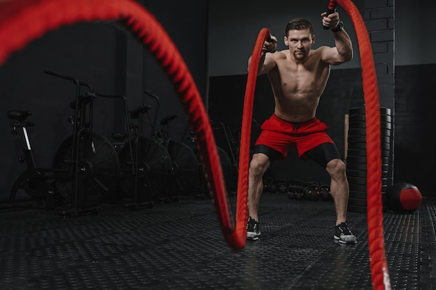 Exercício com cordas de batalha durante o treinamento crossfit na academia. O atleta usa calção vermelho malhando com corda. O conceito de motivação do esporte. Copie o espaço.