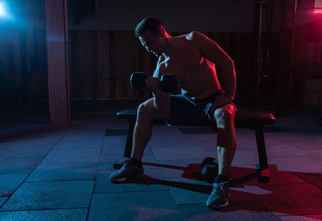 Exercício de bíceps de treinamento de homem musculoso com haltere em luz gradiente vermelha azul no corredor escuro de ajuste cruzado