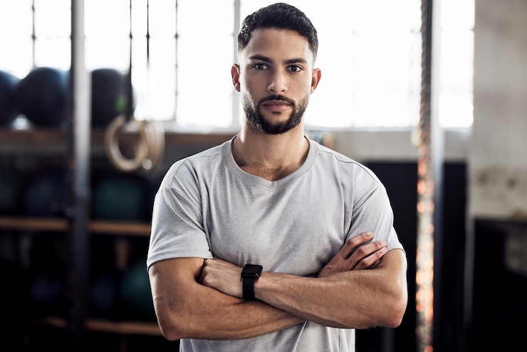 Foto exercício físico e retrato de um homem sério na academia para um treino com foco rosto de atleta masculino ou personal trainer com forte força muscular e motivação com braços cruzados no clube