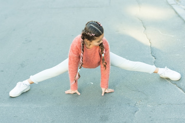 Exercícios de alongamento para prevenir lesões esportivas. Pequena ginasta ao ar livre. Criança pequena faz divisões. Esportes acrobáticos e ginástica. Treinamento físico e esportes infantis. Praticar esportes para ficar em forma.