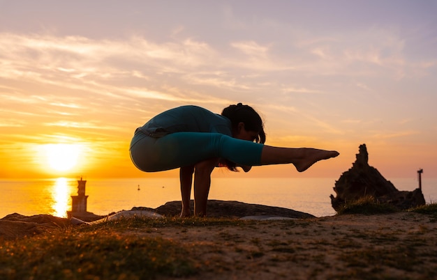 Exercícios de alongamento uma mulher fazendo exercícios de meditação e ioga em uma rocha ao pôr do sol ao lado de um farol no mar vida saudável e naturista