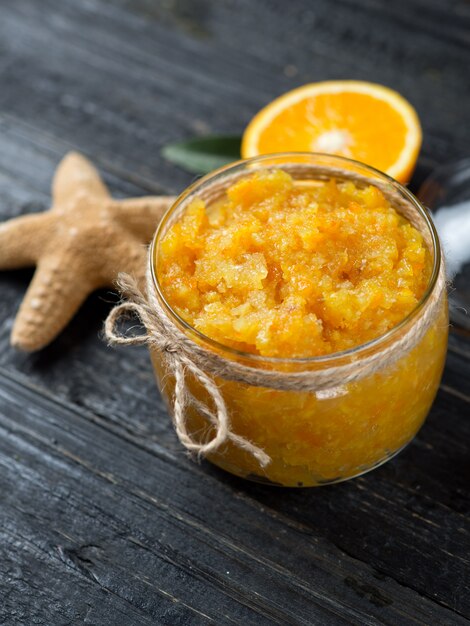 Exfoliante de naranja en un frasco de vidrio sobre una mesa de madera con rodajas de naranja y una estrella de mar