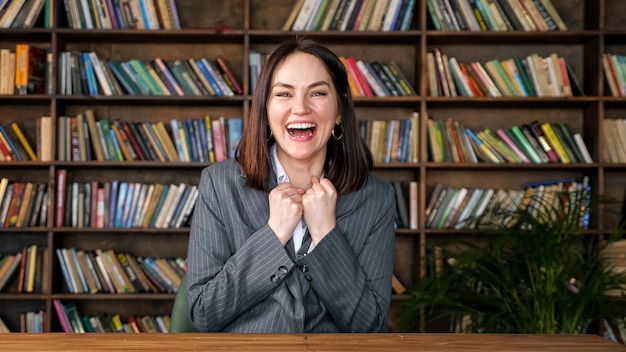 Exitosa empresaria en blusa blanca debajo de la chaqueta gris levanta las manos con felicidad sentado a la mesa contra libros de colores en estantes