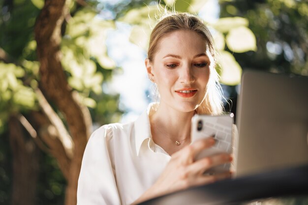 Exitosa empresaria joven sentada en la cafetería al aire libre y con smartphone