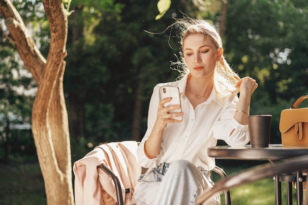 Exitosa empresaria joven sentada en la cafetería al aire libre y con smartphone