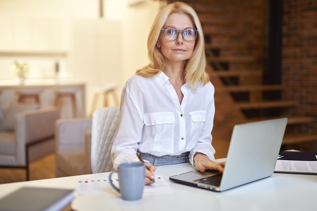 Exitosa empresaria madura en gafas mirando a la cámara con un portátil mientras hace algunos