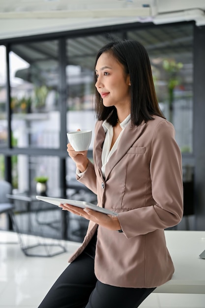 Exitosa mujer de negocios asiática se sienta en la mesa sosteniendo su tableta y taza de café en la oficina