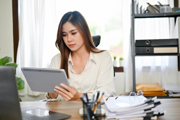 Exitosa mujer de negocios asiática en su escritorio usando una tableta para volver a verificar sus datos financieros