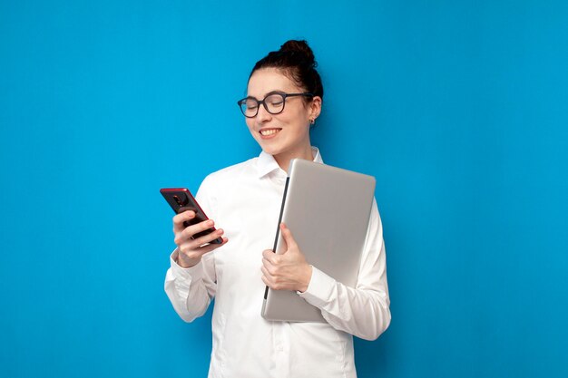 Exitosa mujer de negocios con camisa blanca y anteojos sostiene una computadora portátil y usa un teléfono inteligente