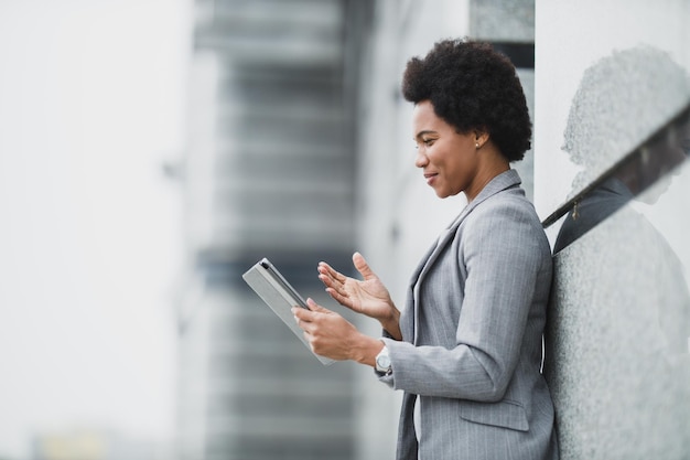 Una exitosa mujer de negocios negra haciendo videollamadas en una tableta digital durante un breve descanso frente a un edificio corporativo.