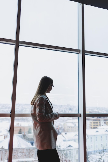 Una exitosa mujer de negocios con pantalones y chaqueta se para y mira por la enorme ventana panorámica del rascacielos del centro de negocios