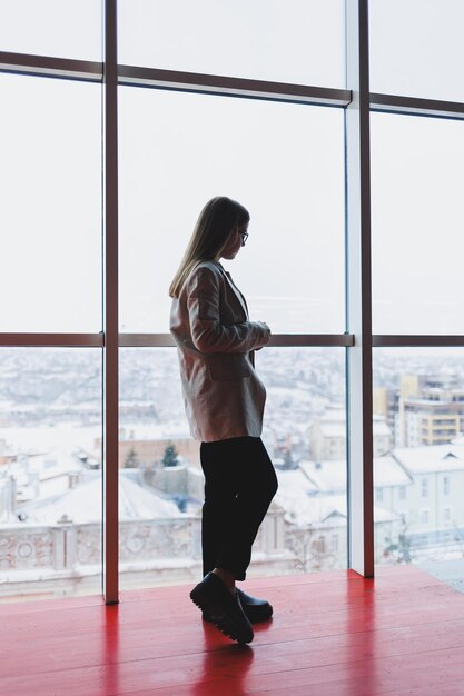 Una exitosa mujer de negocios con pantalones y chaqueta se para y mira por la enorme ventana panorámica del rascacielos del centro de negocios