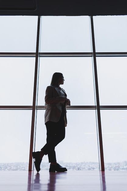 Una exitosa mujer de negocios con pantalones y chaqueta se para y mira por la enorme ventana panorámica del rascacielos del centro de negocios