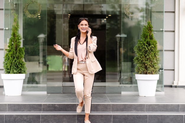 Exitosa mujer de negocios saliendo del edificio de oficinas y conversando por teléfono