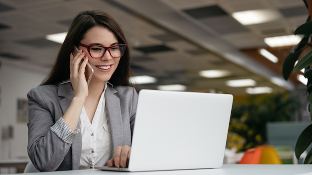 Exitosa mujer de negocios usando una computadora portátil, hablando por teléfono móvil, trabajando en la oficina