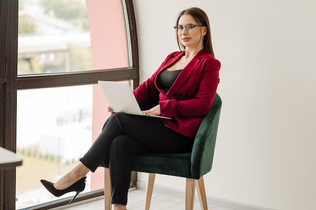 Exitosa mujer de negocios vestida con chaqueta roja está trabajando con una computadora portátil en la oficina