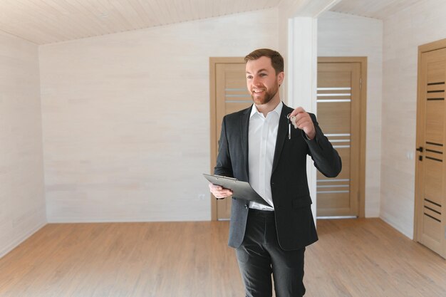 Foto exitoso agente inmobiliario con traje sonríe y ofrece llaves de un nuevo apartamento parado en medio de una habitación que está siendo renovada amplia casa nueva en venta por un corredor profesional de bienes raíces