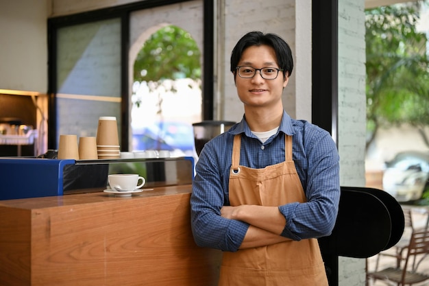 Exitoso y amigable barista asiático o camarero en puestos de delantal con los brazos cruzados en el café