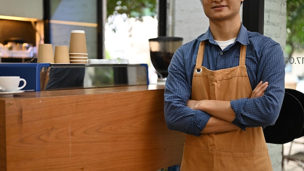 Exitoso barista asiático o camarero en delantal con los brazos cruzados frente al mostrador