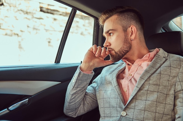 Exitoso y elegante hombre de negocios joven con traje gris y camisa de ping en un asiento trasero en un auto de lujo.