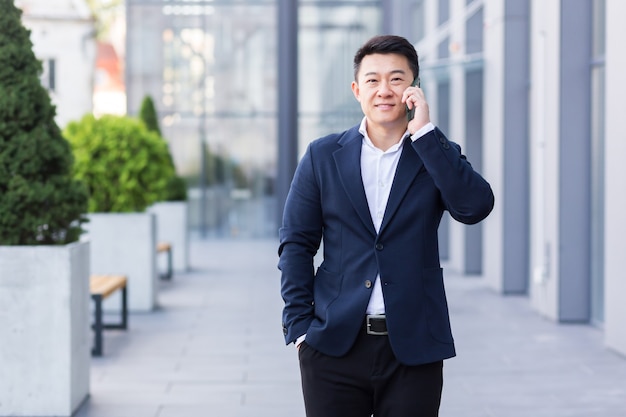 Exitoso empresario asiático empresario hablando por teléfono celular mientras camina cerca de la gran oficina moderna, alegre trabajador sonriente y mirando a la cámara