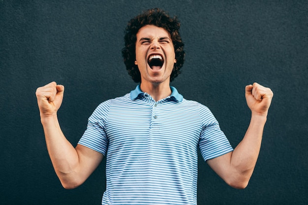 Foto exitoso estudiante o hombre de negocios con cabello rizado con camiseta azul gritando con expresión ganadora puños bombeados posando en la pared blanca concepto de emoción de la gente