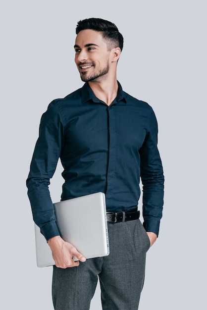 exitoso y guapo. Apuesto joven en camisa que lleva el portátil y apartar la mirada con una sonrisa