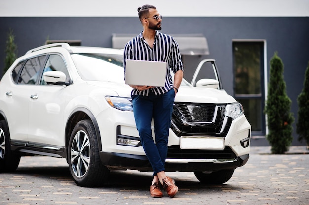 El exitoso hombre árabe vestido con camisa a rayas y gafas de sol posa cerca de su auto blanco con una computadora portátil en la mano.