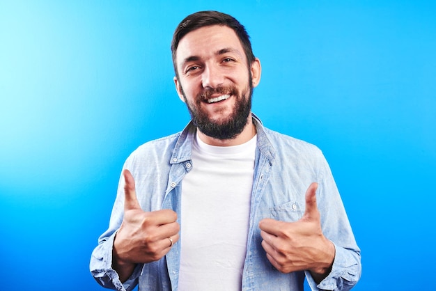 Exitoso hombre barbudo caucásico guapo muestra los pulgares hacia arriba y sonrisas con dientes en el fondo azul del estudio aislado