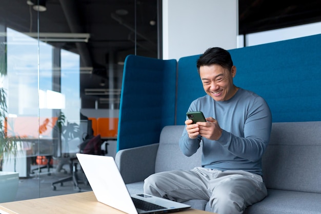 El exitoso hombre de negocios asiático celebra la victoria, mira el teléfono feliz y se regocija sentado en la oficina moderna