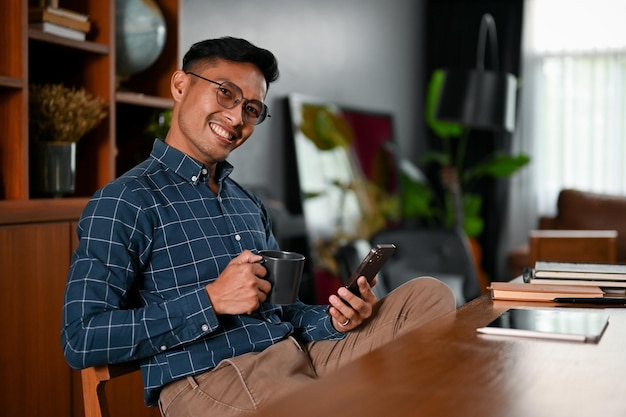 Exitoso hombre de negocios asiático tomando café y usando su teléfono inteligente en su escritorio