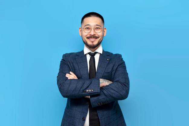 exitoso hombre de negocios asiático con traje y gafas está de pie con los brazos cruzados sobre fondo azul