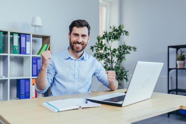 Exitoso hombre de negocios con barba mira a la cámara y sonríe celebra el triunfo trabaja en una laptop en la oficina