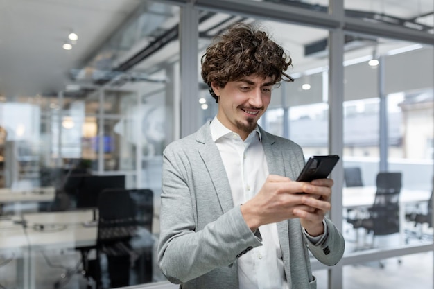 Exitoso hombre de negocios con barba de pie cerca de la ventana hombre usando teléfono feliz jefe escribiendo mensaje y