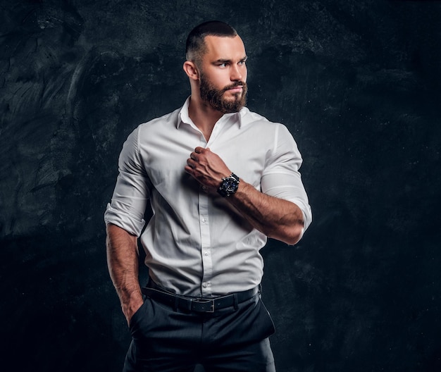 Un exitoso hombre de negocios barbudo con camisa blanca posa en un estudio fotográfico oscuro.