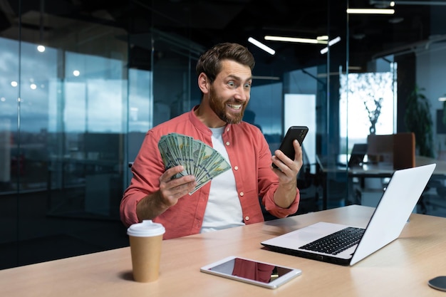 Foto exitoso hombre de negocios con camisa roja celebrando la victoria y el buen logro con teléfono y dinero