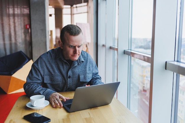 El exitoso hombre de negocios feliz está sentado en una mesa en un café sosteniendo una taza de café y usando una computadora portátil