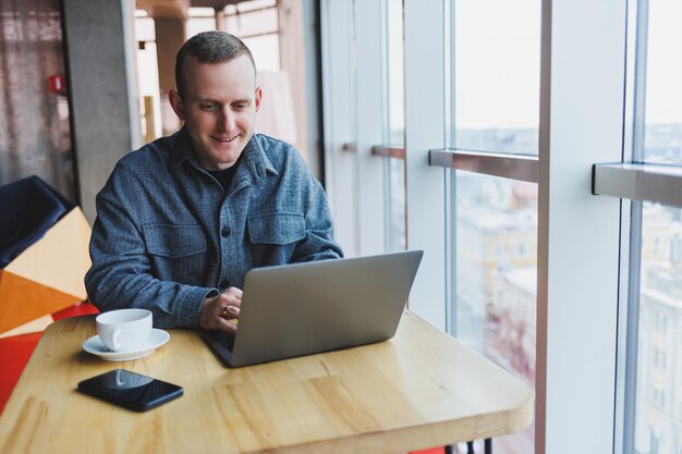 El exitoso hombre de negocios feliz está sentado en una mesa en un café, sosteniendo una taza de café y usando una computadora portátil.