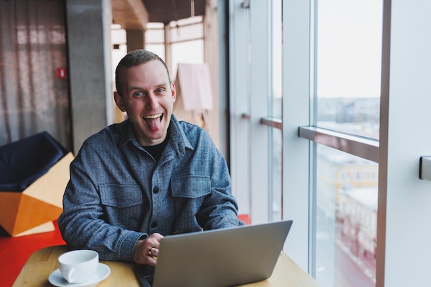 El exitoso hombre de negocios feliz está sentado en una mesa en un café sosteniendo una taza de café y usando una computadora portátil