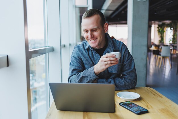 El exitoso hombre de negocios feliz está sentado en una mesa en un café sosteniendo una taza de café y usando una computadora portátil