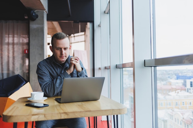 El exitoso hombre de negocios feliz está sentado en una mesa en un café sosteniendo una taza de café y usando una computadora portátil