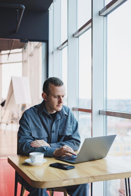 Foto el exitoso hombre de negocios feliz está sentado en una mesa en un café, sosteniendo una taza de café y usando una computadora portátil.