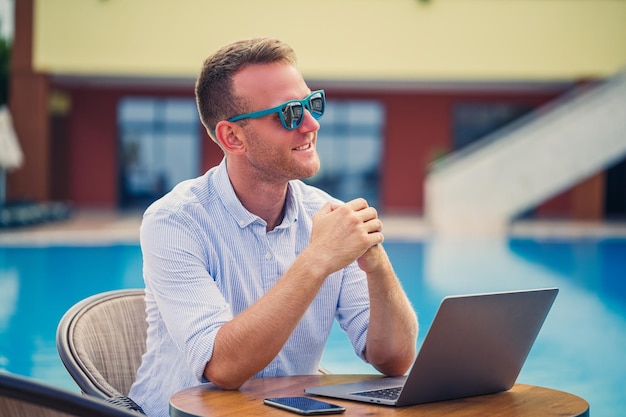 Exitoso hombre de negocios guapo con gafas de sol trabaja en una computadora portátil sentada cerca de la piscina Trabajo remoto Freelancer