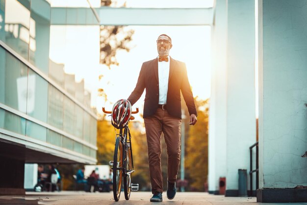 Un exitoso hombre de negocios de mediana edad con una bicicleta a su lado camina frente al distrito de oficinas y va a la oficina.