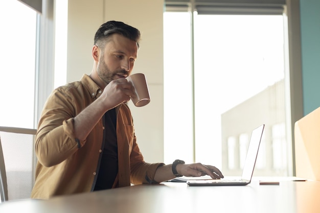 Exitoso hombre de negocios que trabaja en línea en una computadora portátil tomando café en la oficina