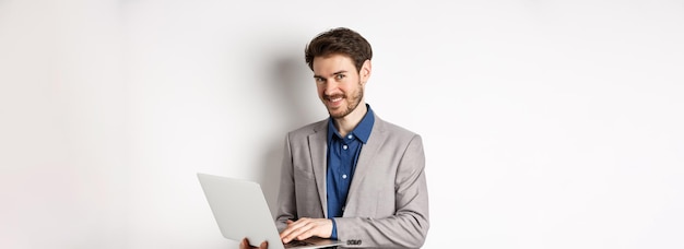 Exitoso hombre de negocios sonriente trabajando en una laptop y mirando feliz a la cámara de pie con traje gris o