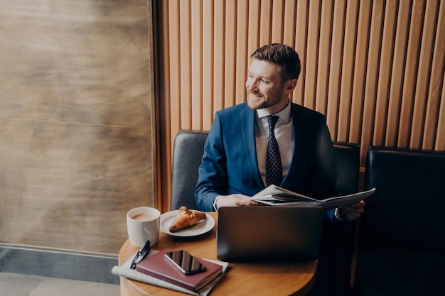 Exitoso hombre de negocios sonriente en traje formal azul mirando afuera de la ventana, sosteniendo el periódico mientras está sentado en la cafetería con computadora portátil después de pedir café con croissant