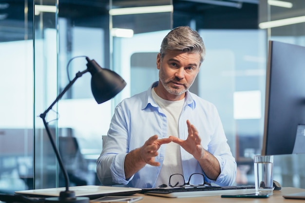 El exitoso hombre de negocios trabaja en la computadora, se sienta en la mesa y mira a la cámara, se comunica, explica y gesticula