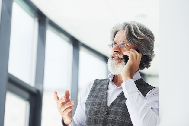Exitoso hombre de negocios usa teléfono Senior hombre moderno con estilo con cabello gris y barba en el interior
