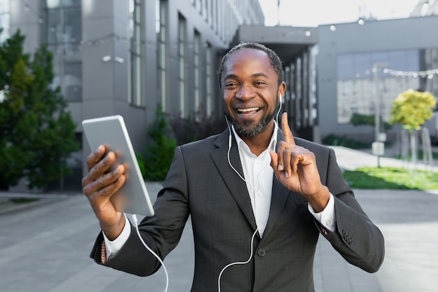 Exitoso jefe afroamericano fuera del edificio de oficinas con tableta y auriculares hablando